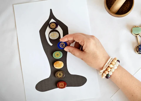 A hand placing painted chakra stones along a seated meditating figure, with mala beads on the wrist and a singing bowl beside it.